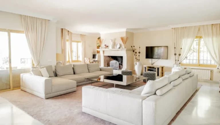 A bright living room with two cream sofas, a stone fireplace and tall curtained windows